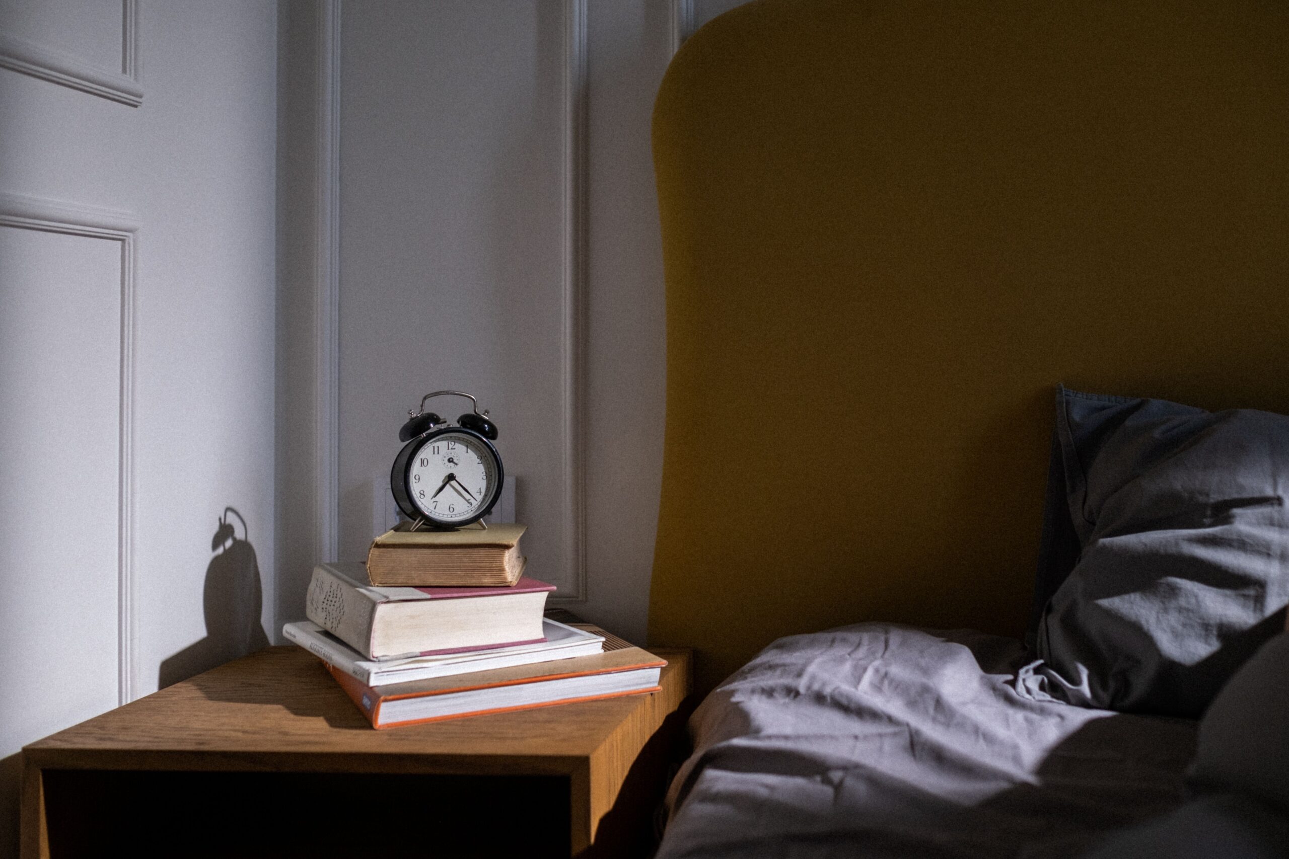 How To Choose The Perfect Nightstands For Your Bedroom A cozy bedroom setting with books and an alarm clock on a nightstand next to a bed.