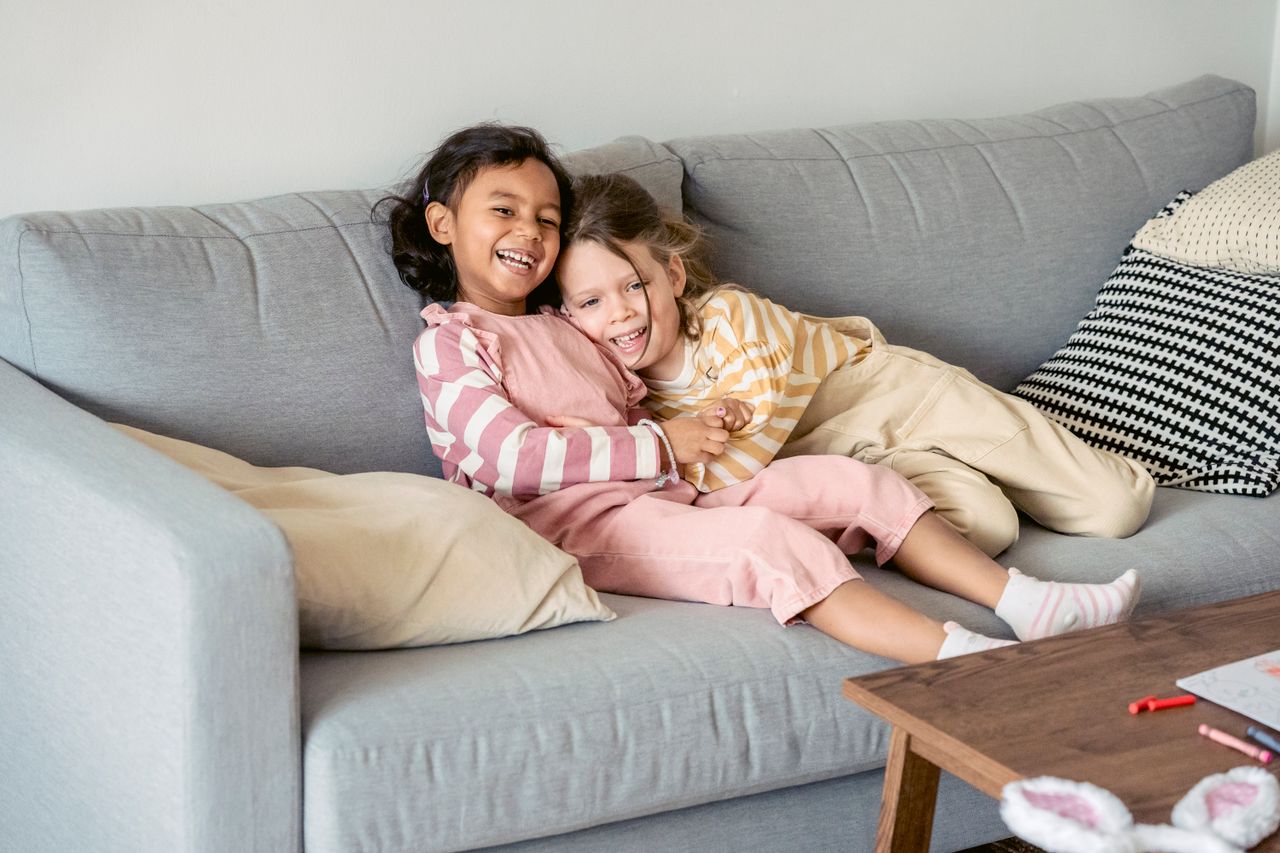 Cheerful multiethnic girlfriends wearing similar outfits cuddling on comfortable sofa at home near table with chalk pencils